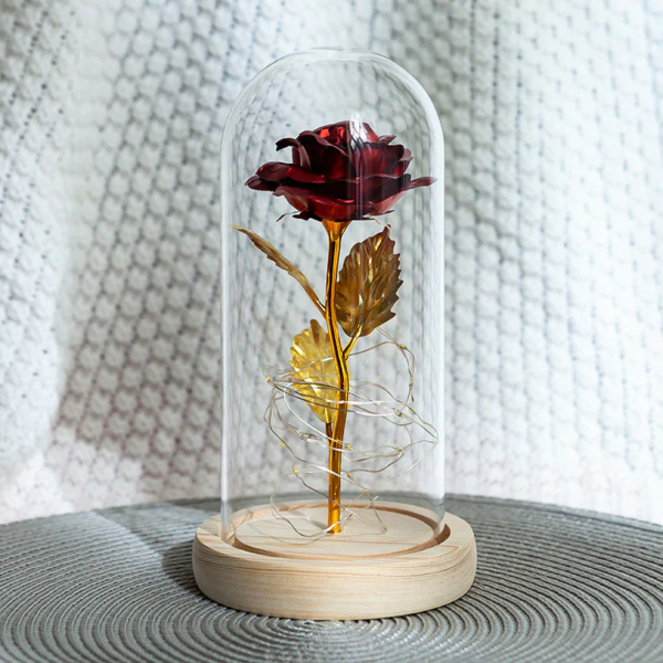 A preserved red rose with gold-plated stem and leaves, encased in a glass dome with warm LED lights, resting on a wooden base.