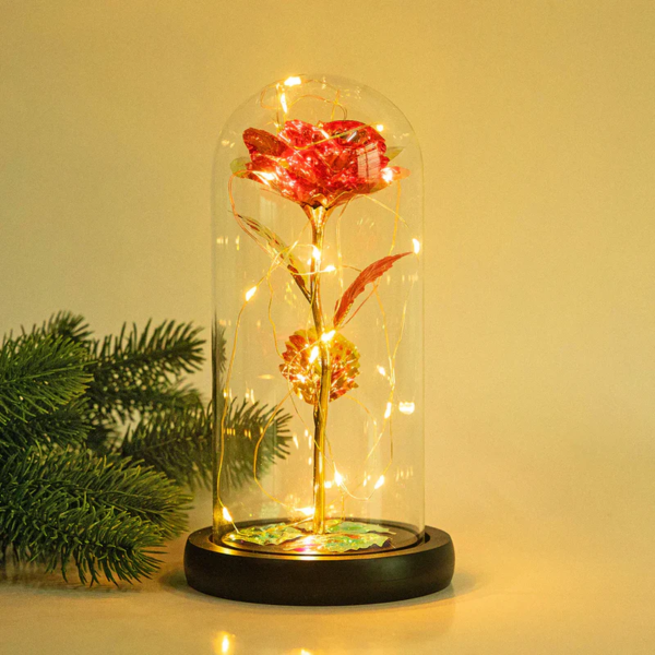 A preserved red rose with gold-plated stem and leaves, encased in a glass dome with warm LED lights, resting on a wooden base.