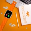 An Ultra 2 Smart Watch 7 with an orange sport band displaying an incoming call notification, placed on a desk with a laptop and notebook.