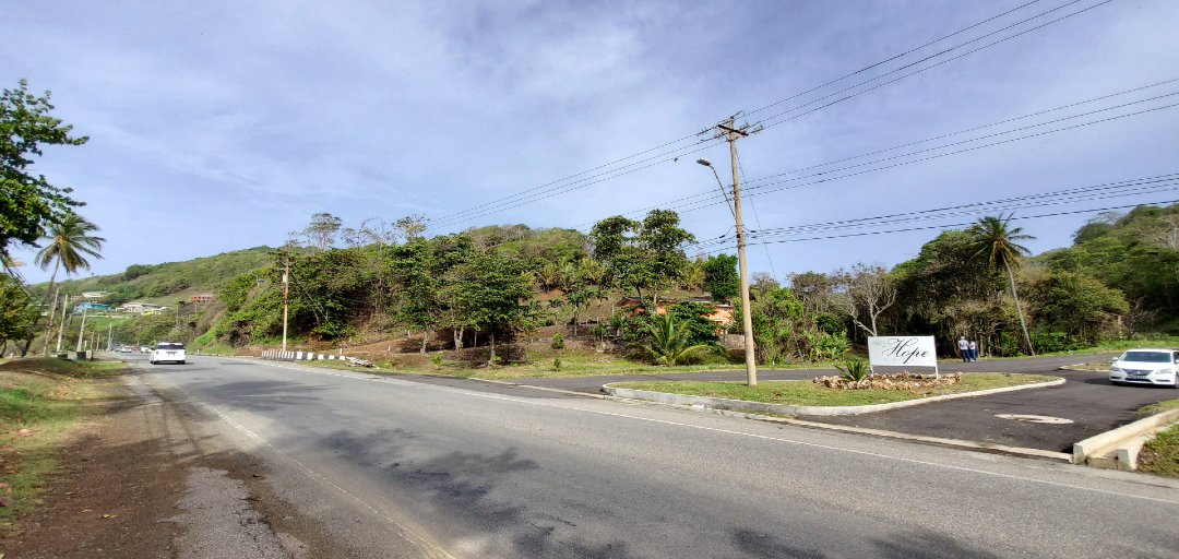 A roadside view of the entrance to Hope Estate, Tobago, with lush greenery and a glimpse of the community beyond.