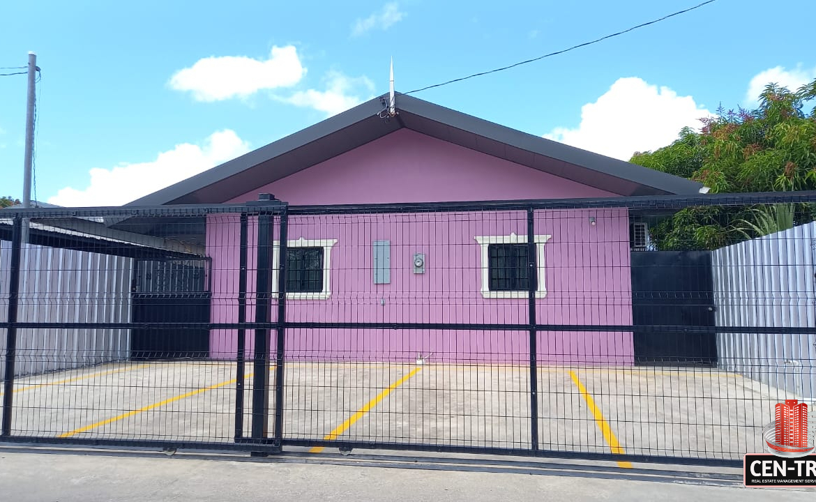 Front view of a pink one-story apartment building with a black metal security gate and a paved parking area of this lovely apartment for rent