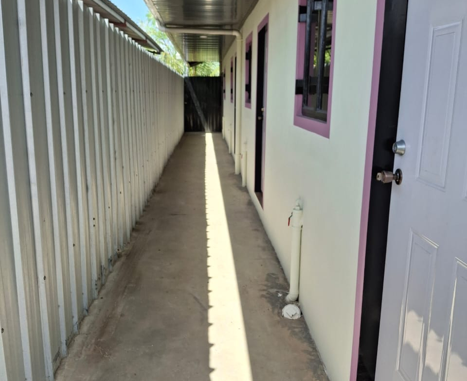 Exterior view of a narrow paved walkway alongside an apartment building with light-colored walls, purple trim around the windows, and white doors. A gray metal fence runs along one side of the walkway, and a black security gate is visible in the distance. The sky is blue with scattered clouds.