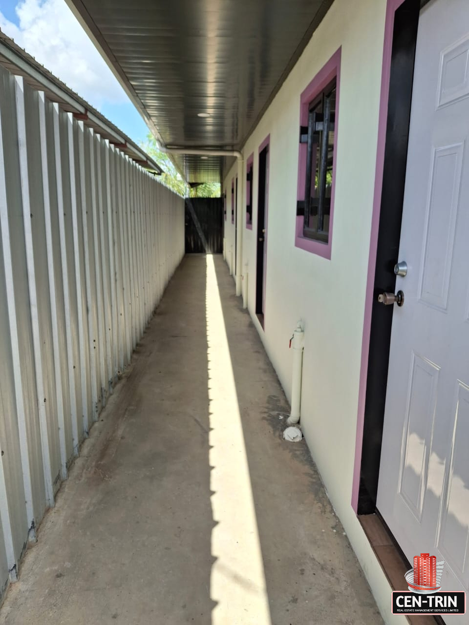 Exterior view of a narrow paved walkway alongside an apartment building with light-colored walls, purple trim around the windows, and white doors. A gray metal fence runs along one side of the walkway, and a black security gate is visible in the distance. The sky is blue with scattered clouds.