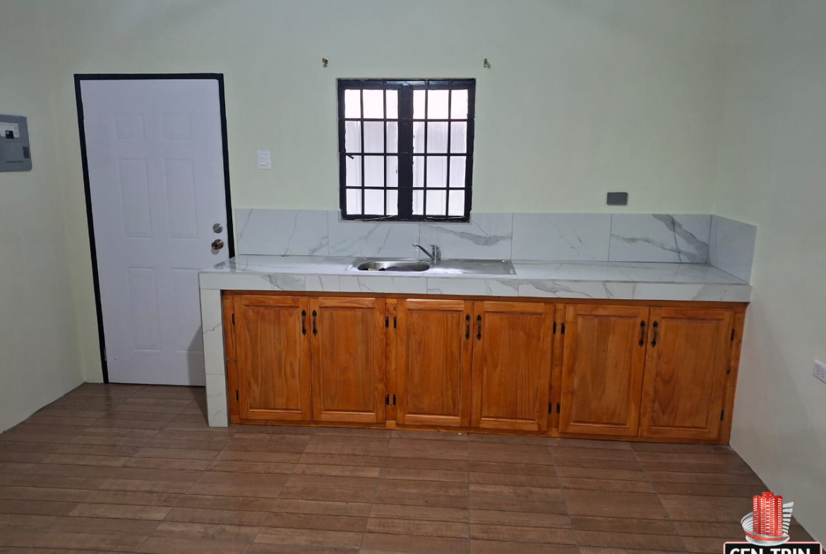 Interior view of an apartment kitchen featuring wooden cabinets, a marble-like countertop, a stainless steel sink, a window with bars, and a white door with a black frame.