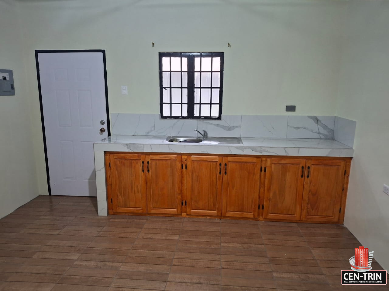 Interior view of an apartment kitchen featuring wooden cabinets, a marble-like countertop, a stainless steel sink, a window with bars, and a white door with a black frame.