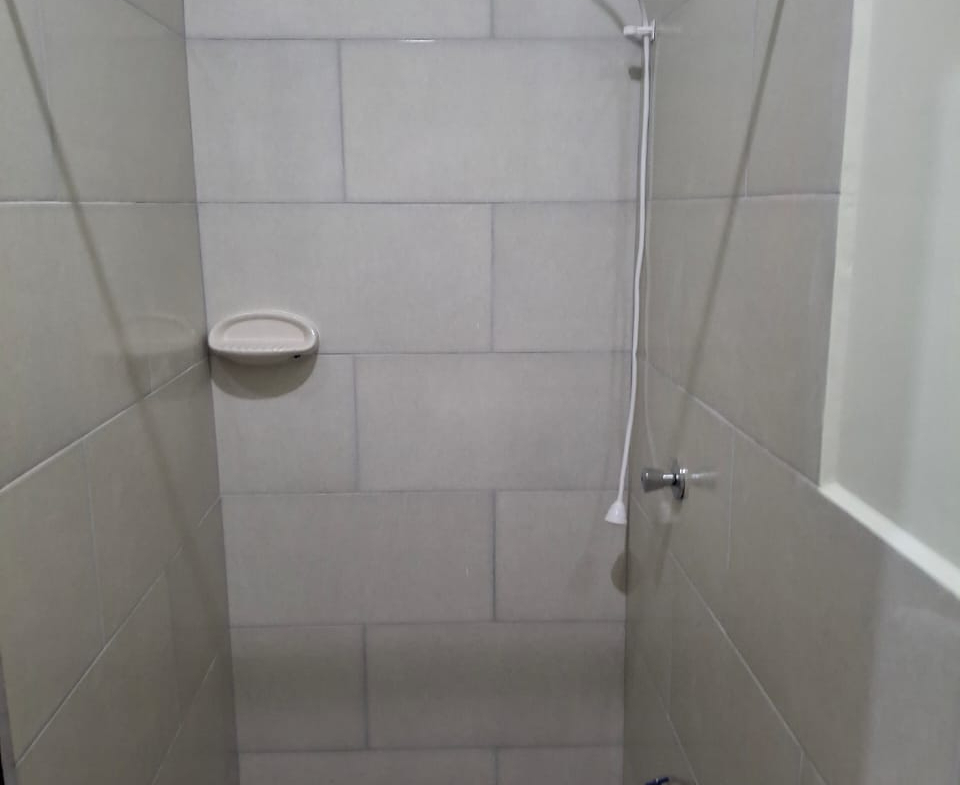 Close-up interior view of an apartment bathroom featuring a shower area with light gray tiled walls, a shower head, a soap dish, and a patterned gray and white tiled floor. A white sink is partially visible in the foreground.