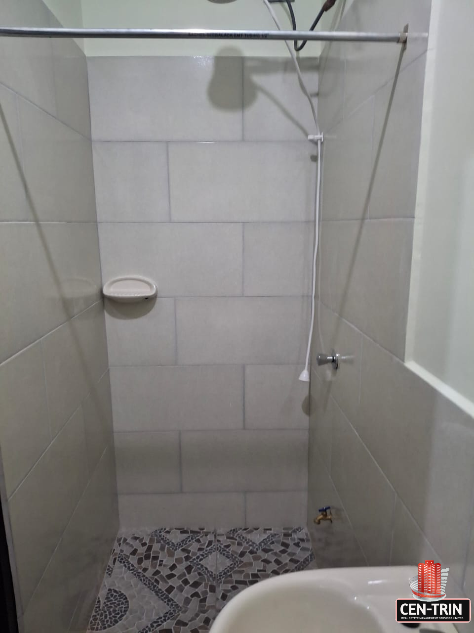 Close-up interior view of an apartment bathroom featuring a shower area with light gray tiled walls, a shower head, a soap dish, and a patterned gray and white tiled floor. A white sink is partially visible in the foreground.