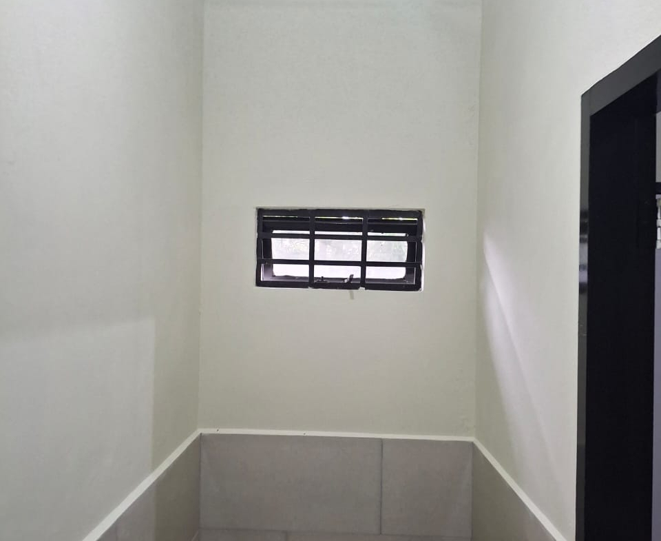 Interior view of an apartment bathroom featuring a toilet, a small barred window, light gray tiled walls, a decorative patterned ceiling, and a metal shower rod.