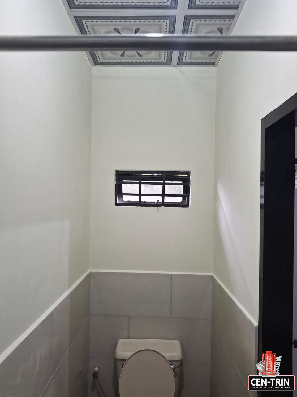 Interior view of an apartment bathroom featuring a toilet, a small barred window, light gray tiled walls, a decorative patterned ceiling, and a metal shower rod.