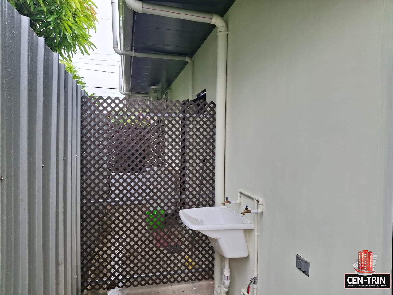 Exterior view of a section of an apartment building with light green walls, showing a white utility sink mounted on the wall, white PVC pipes, a gray metal fence, and a brown lattice fence. Green foliage is visible at the top.