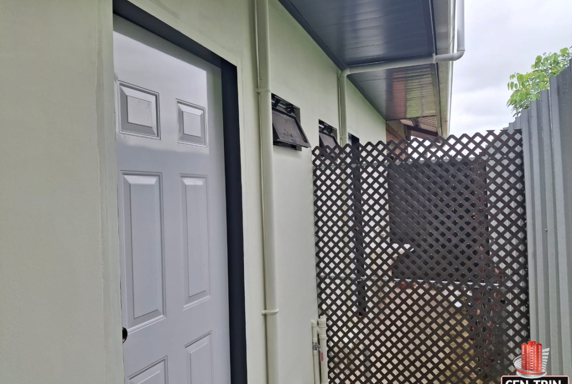 Exterior view of an apartment building with light green walls, featuring a partially open white door with a black frame, white PVC pipes, a small window, a gray metal fence, and a brown lattice fence.