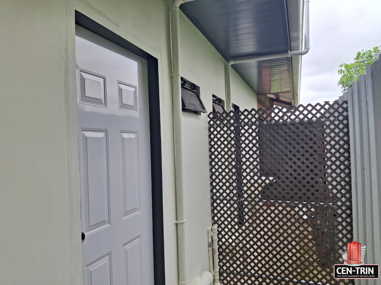 Exterior view of an apartment building with light green walls, featuring a partially open white door with a black frame, white PVC pipes, a small window, a gray metal fence, and a brown lattice fence.