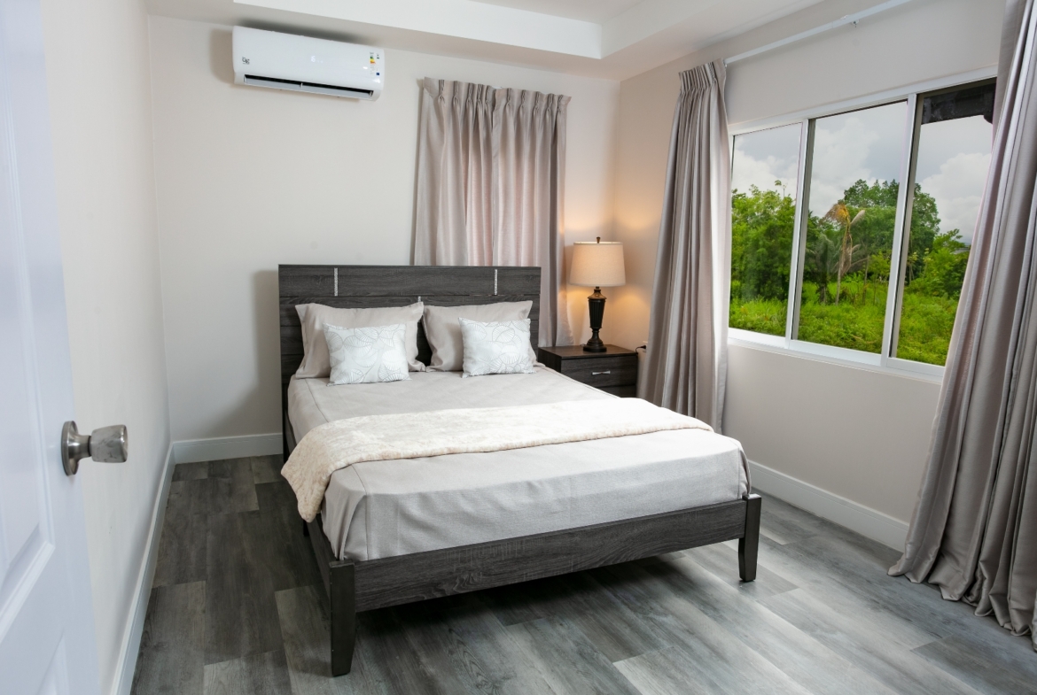 Bedroom at Roystonia Mews featuring a bed with pillows, curtains, a window with an outdoor view, and gray plank flooring.