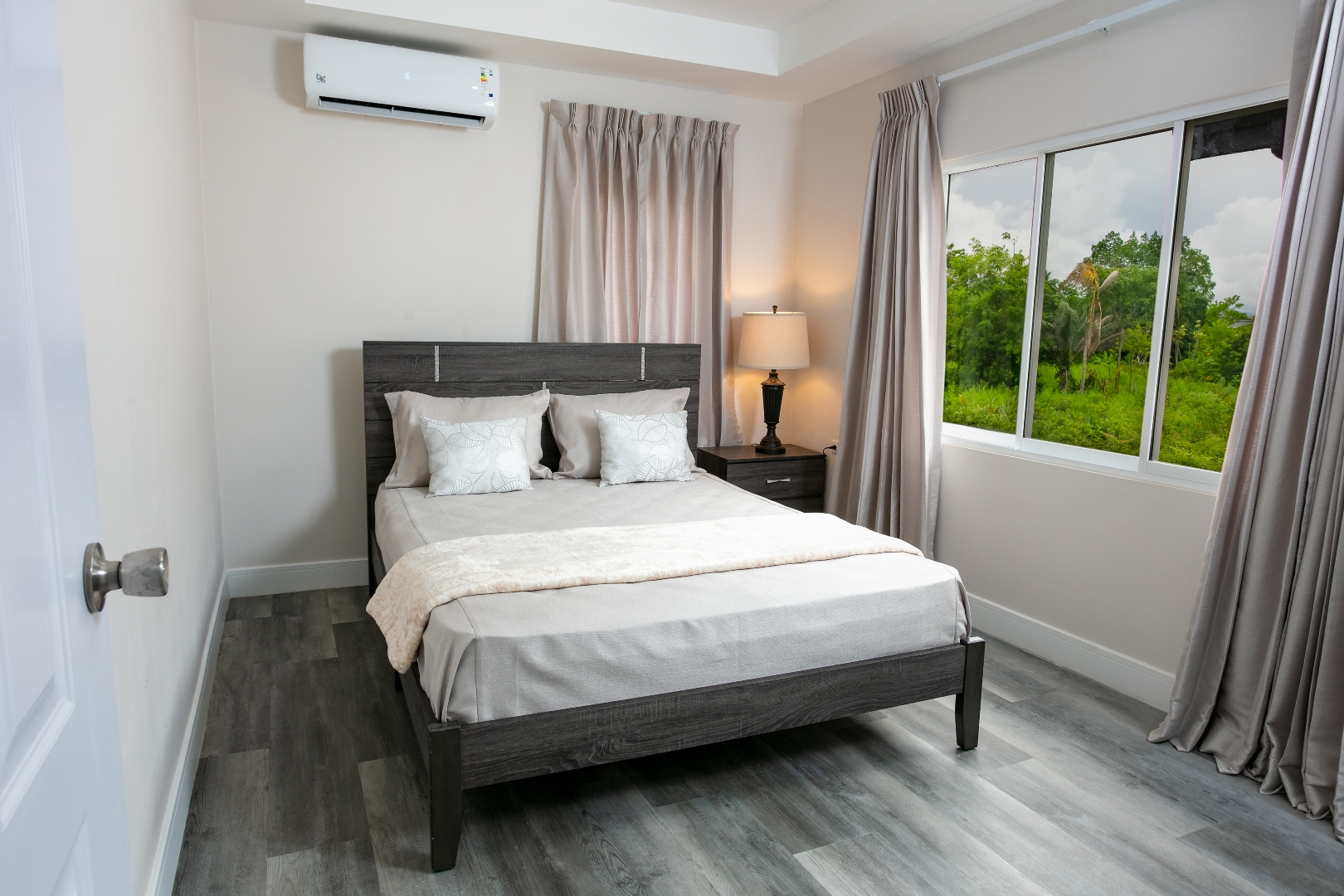 Bedroom at Roystonia Mews featuring a bed with pillows, curtains, a window with an outdoor view, and gray plank flooring.