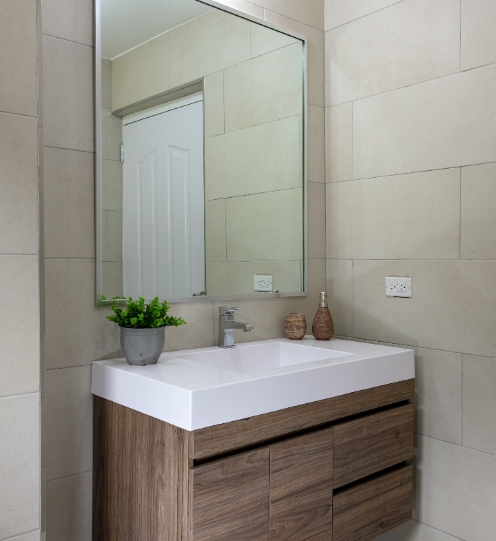 Bathroom vanity at Roystonia Mews featuring a white countertop, floating wooden cabinet, large mirror, tiled walls, and a small potted plant.