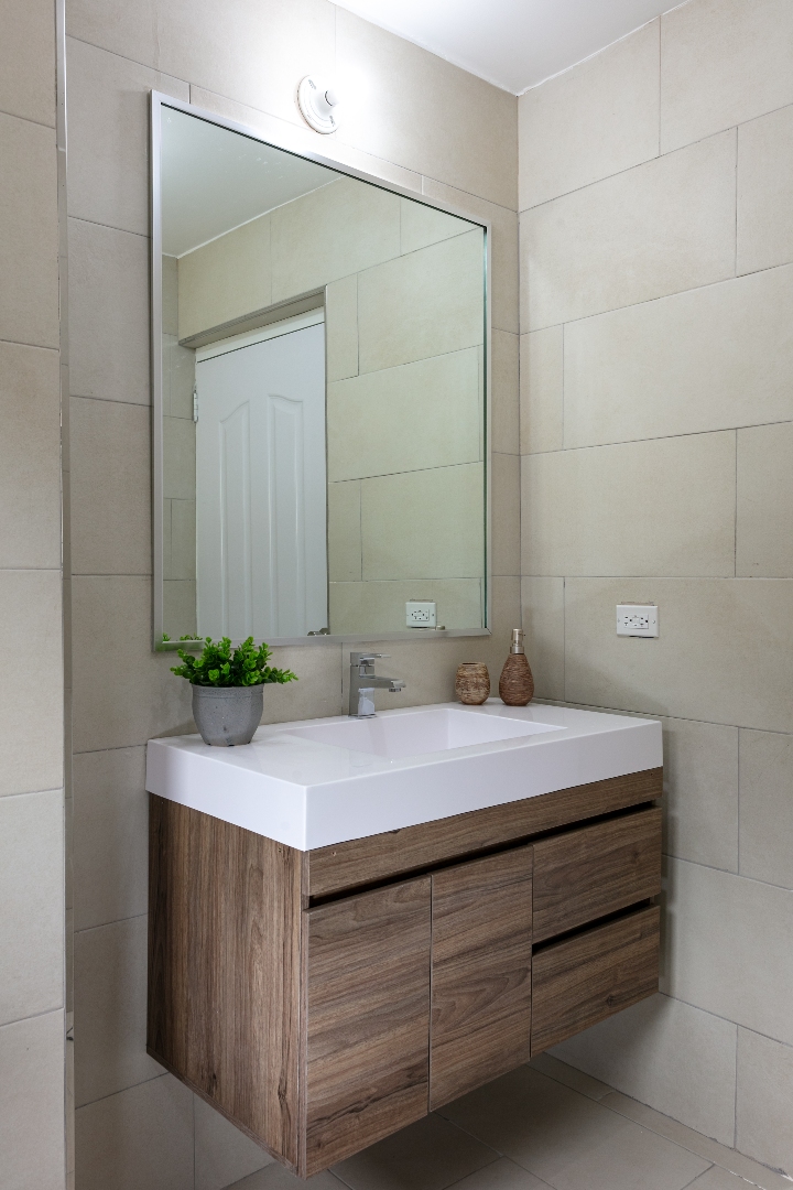 Bathroom vanity at Roystonia Mews featuring a white countertop, floating wooden cabinet, large mirror, tiled walls, and a small potted plant.