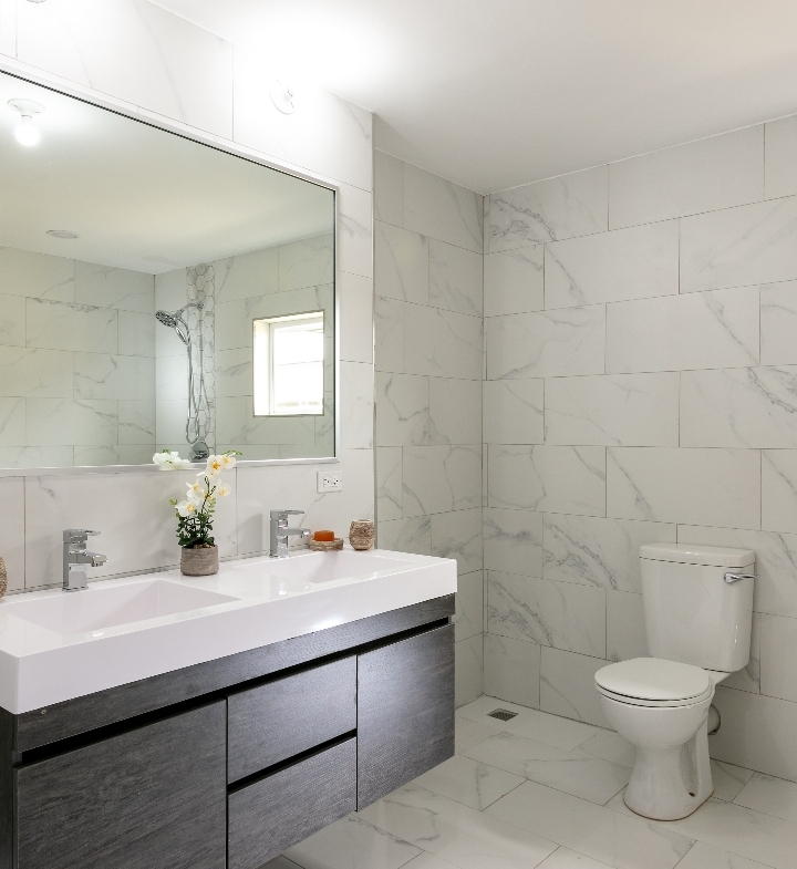 Spacious bathroom at Roystonia Mews featuring a double vanity, large mirror, tiled walls and floors, and a toilet.