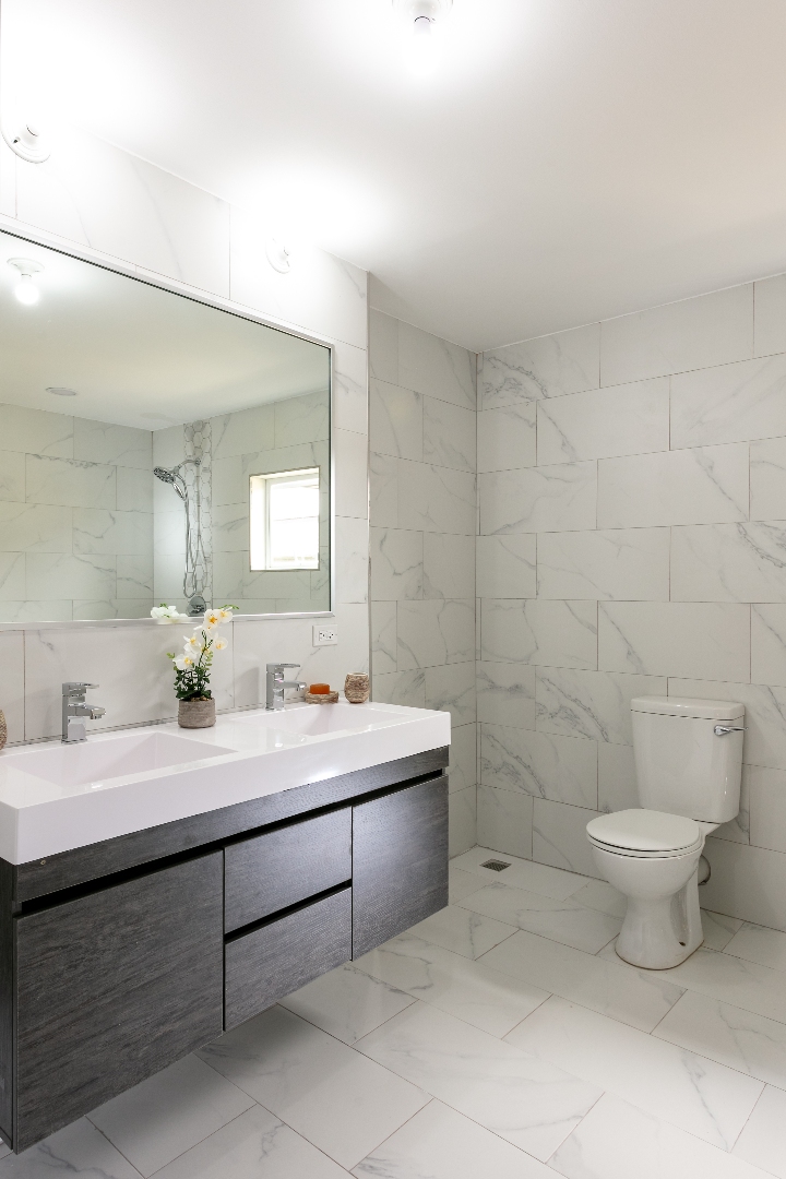 Spacious bathroom at Roystonia Mews featuring a double vanity, large mirror, tiled walls and floors, and a toilet.