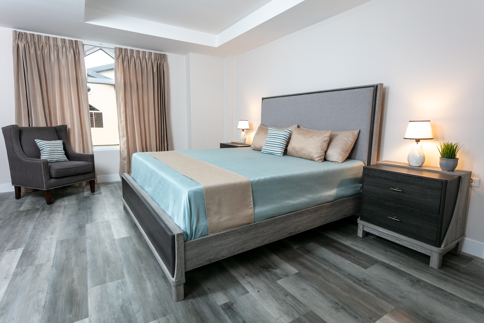 Master bedroom at Roystonia Mews featuring a bed with pillows, an armchair, curtains, a tray ceiling, and gray plank flooring.