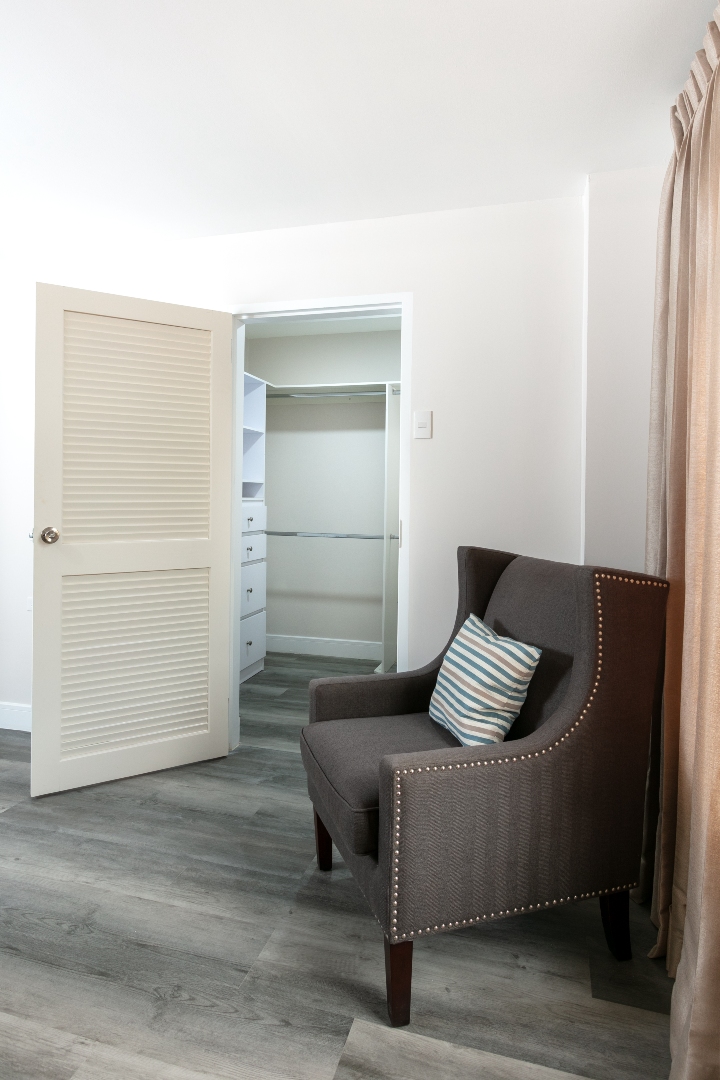 Master bedroom at Roystonia Mews featuring a wing chair, open walk-in closet with built-in shelving, and gray plank flooring.