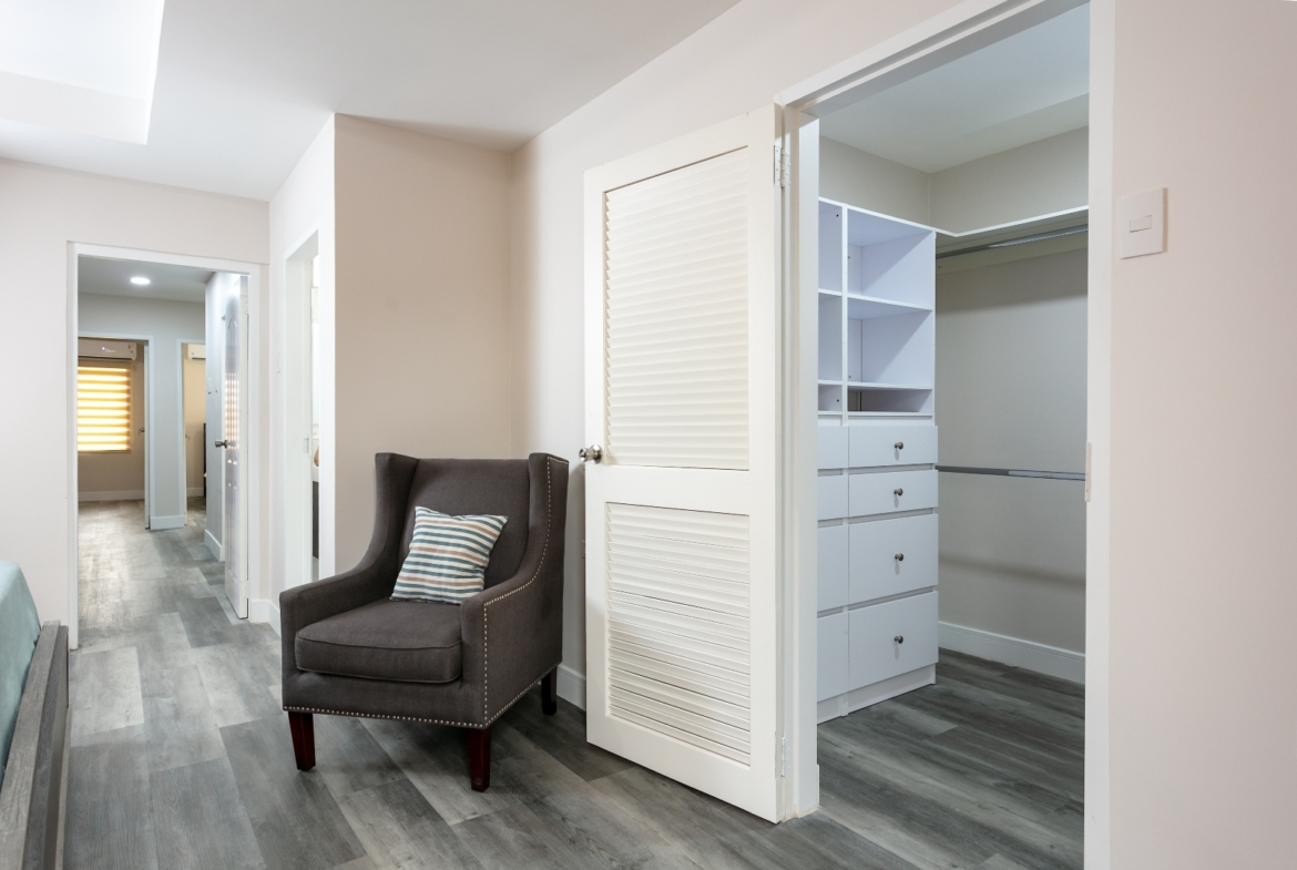 Master suite hallway at Roystonia Mews featuring a wing chair, open walk-in closet with built-in shelves and drawers, and gray plank flooring.