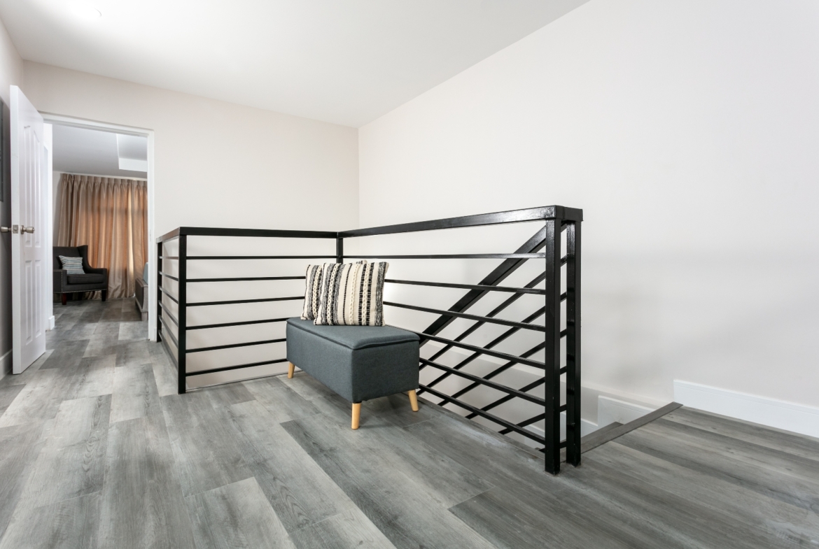 Upstairs hallway at Roystonia Mews featuring a modern black railing, gray plank flooring, and a small bench with a striped cushion.