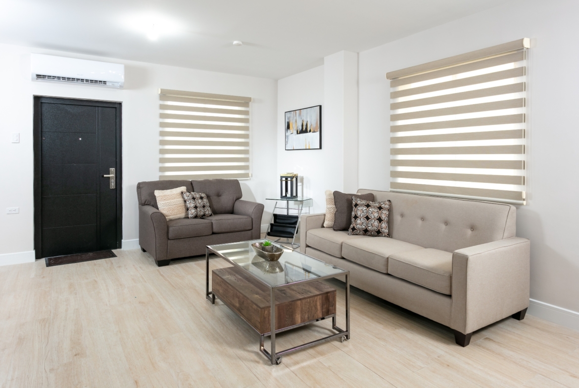 Interior view of a living room in Roystonia Mews, showing a dark front door, air conditioning unit, modern furniture, and large windows with blinds.