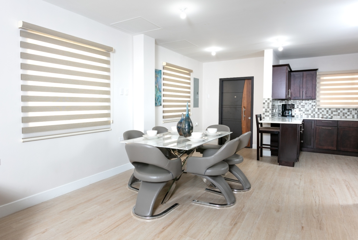 Dining area and kitchen view in Roystonia Mews, featuring a modern dining set, kitchen cabinets, and large windows with blinds.