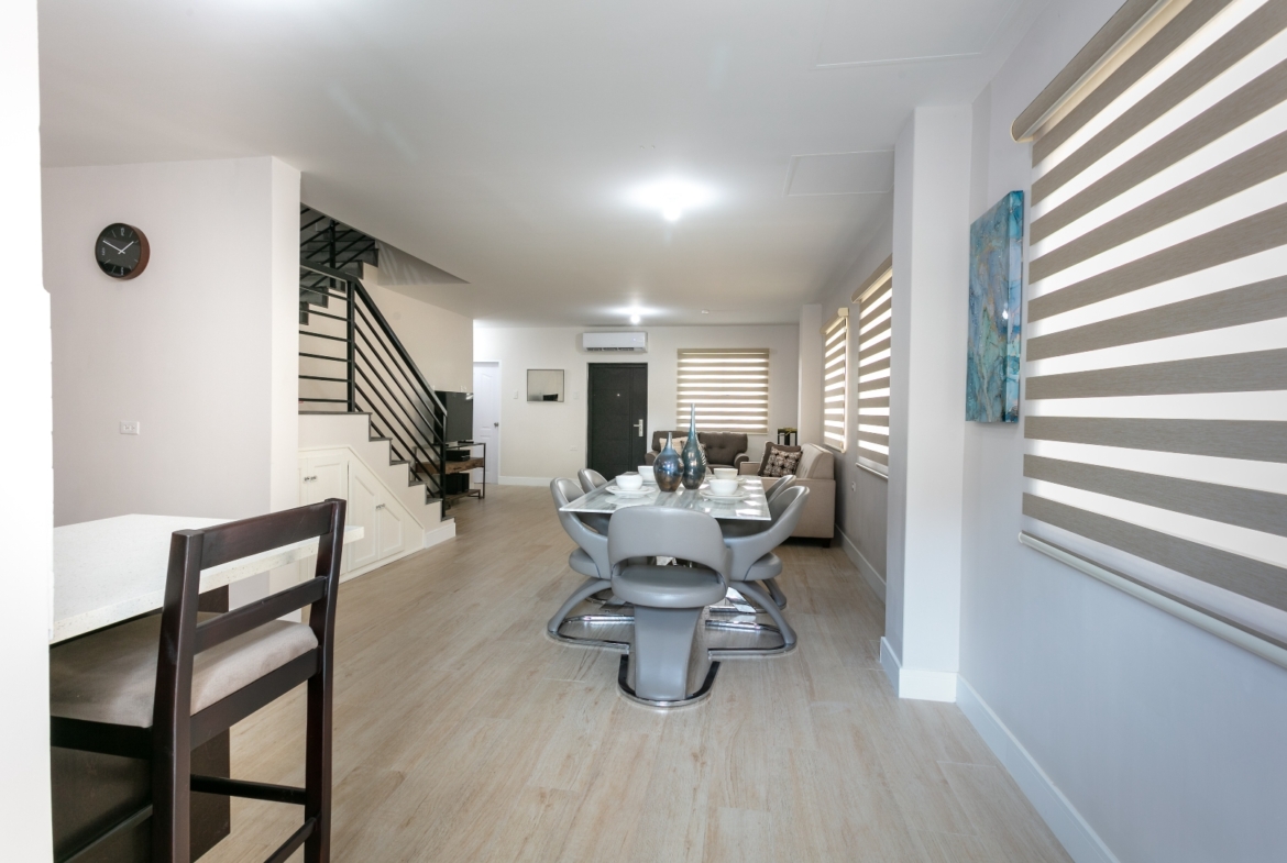 Wide interior view of Roystonia Mews, showing open concept living, dining area, staircase, and natural light from windows with blinds.