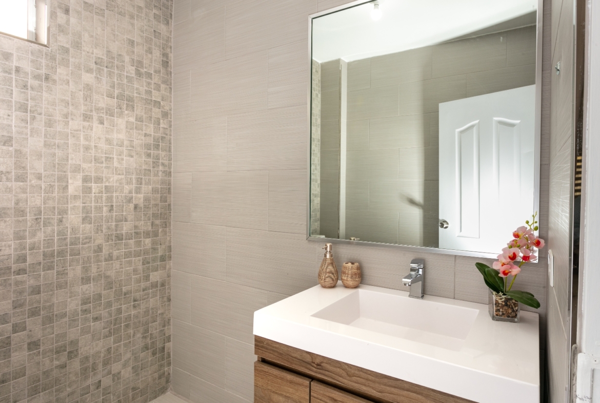 Modern bathroom at Roystonia Mews featuring a vanity with a white countertop and wooden cabinets, a large mirror, tiled walls, and a small window.