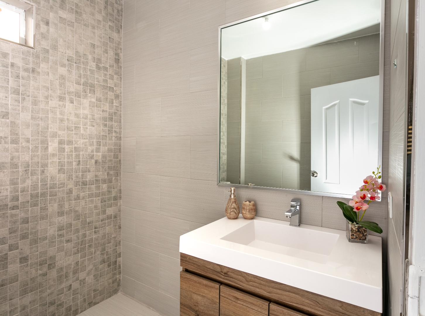 Modern bathroom at Roystonia Mews featuring a vanity with a white countertop and wooden cabinets, a large mirror, tiled walls, and a small window.