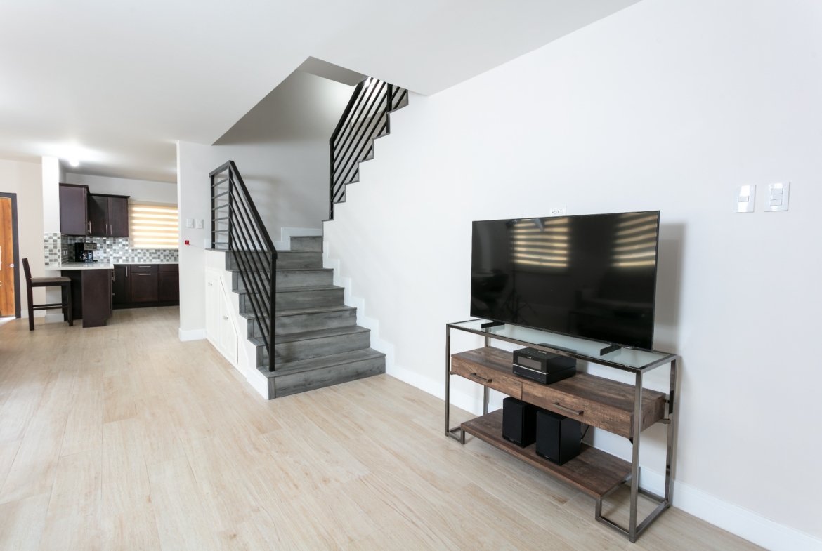 Living area at Roystonia Mews featuring a modern staircase with black railings, a television on a stand, and a view into the kitchen.