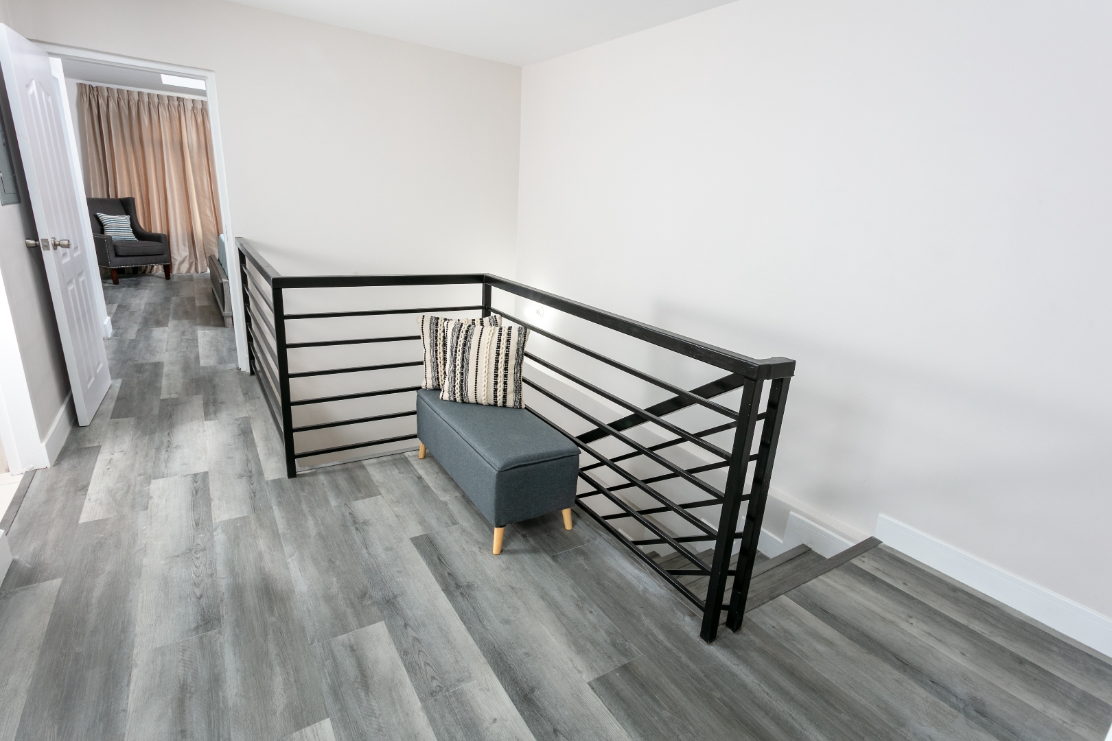 Upstairs hallway at Roystonia Mews featuring a modern staircase railing, gray plank flooring, a small bench, and a doorway leading to a room.