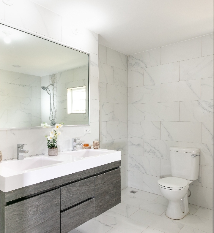 Spacious bathroom at Roystonia Mews featuring a double vanity with a gray floating cabinet, large mirror, white marble-like tile walls and floors, and a toilet.