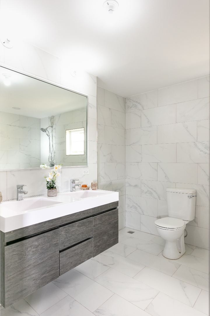 Spacious bathroom at Roystonia Mews featuring a double vanity with a gray floating cabinet, large mirror, white marble-like tile walls and floors, and a toilet.