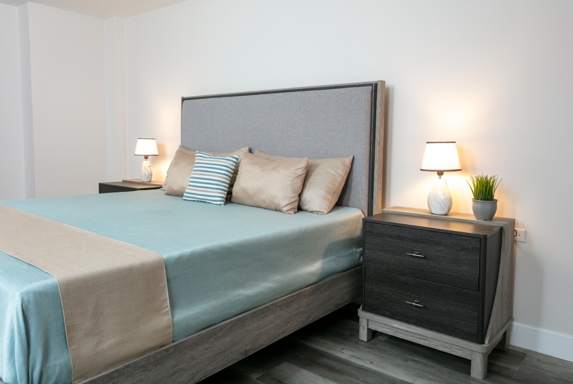 Master bedroom at Roystonia Mews featuring a bed with an upholstered gray headboard, decorative pillows, matching nightstands, and gray plank flooring.